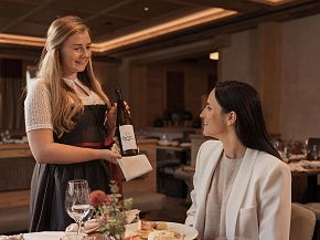 A smiling waitress presents a bottle of wine to a seated woman in a cozy restaurant setting with wooden decor and elegantly set tables.
