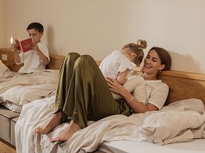 Eine Frau entspannt mit zwei Kindern in einem gemütlichen Hotelzimmer. Ein Kind liest ein Buch, das andere kuschelt mit der Mutter auf dem Bett.