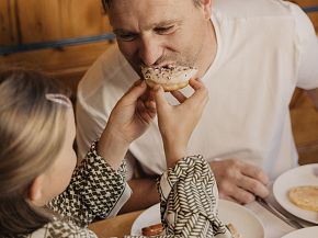 A father and his child share a tender moment during breakfast as the child feeds him a pastry. They sit at a table, smiling in a warm, wooden setting.
