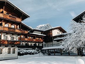 Winterliche Ansicht eines traditionellen alpinen Hotels mit verschneiten Dächern und Bäumen. Berggipfel im Hintergrund bei klarem Himmel.
