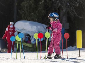 A child in pink ski gear practices skiing through colorful poles on a snowy slope, with an instructor in red in the background.