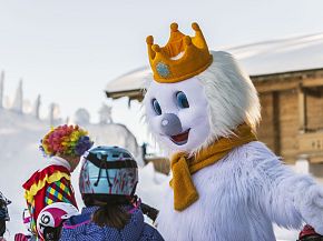 A fluffy white mascot with a crown and scarf interacts with children in winter clothing in a snowy outdoor setting near a wooden chalet.
