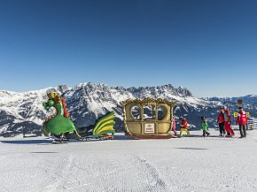A family enjoys a scenic winter day on a snowy mountain with a dragon-themed sleigh. Majestic alpine peaks are visible under clear blue skies.