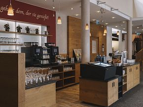 Moderner Hotel-Speiseraum mit Holzdekor und Beleuchtung. Kaffee- und Getränkestation im Vordergrund, im Hintergrund Gläserregale und rote Wand mit Zitat.