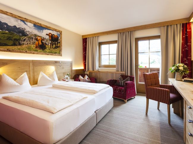 A cozy hotel room with a large bed, warm lighting, and a landscape painting above the headboard. Wooden furniture and mountain views enhance the ambiance.