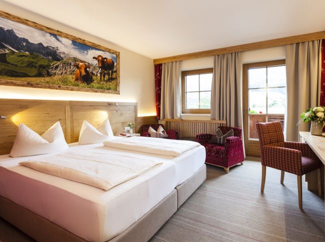 Gemütliches Hotelzimmer mit Doppelbett, Holzverkleidung, Bergbild an der Wand, Sesseln und Fenster mit Vorhängen. Helles, einladendes Ambiente.