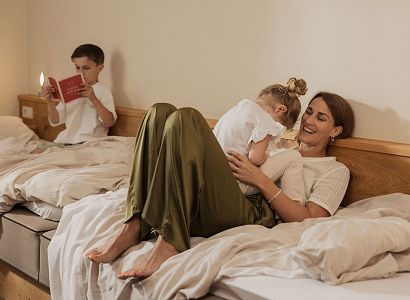 Eine Mutter kuschelt mit ihrer Tochter auf einem Bett, während ein Junge auf dem anderen Bett ein Buch liest. Gemütliche und entspannte Atmosphäre im Zimmer.