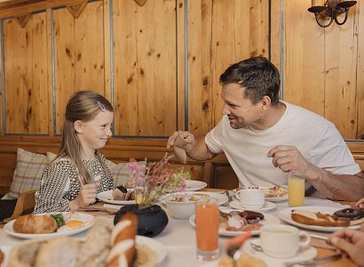Vater und Tochter genießen ein Frühstück in einer gemütlichen Holzstube. Der Tisch ist reich gedeckt mit Brot, Gebäck und frischem Orangensaft.