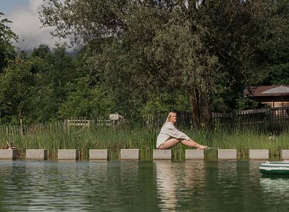 Frau sitzt entspannt am Ufer eines Naturpools, umgeben von grüner Vegetation und einem ruhigen, klaren Himmel im Hintergrund.
